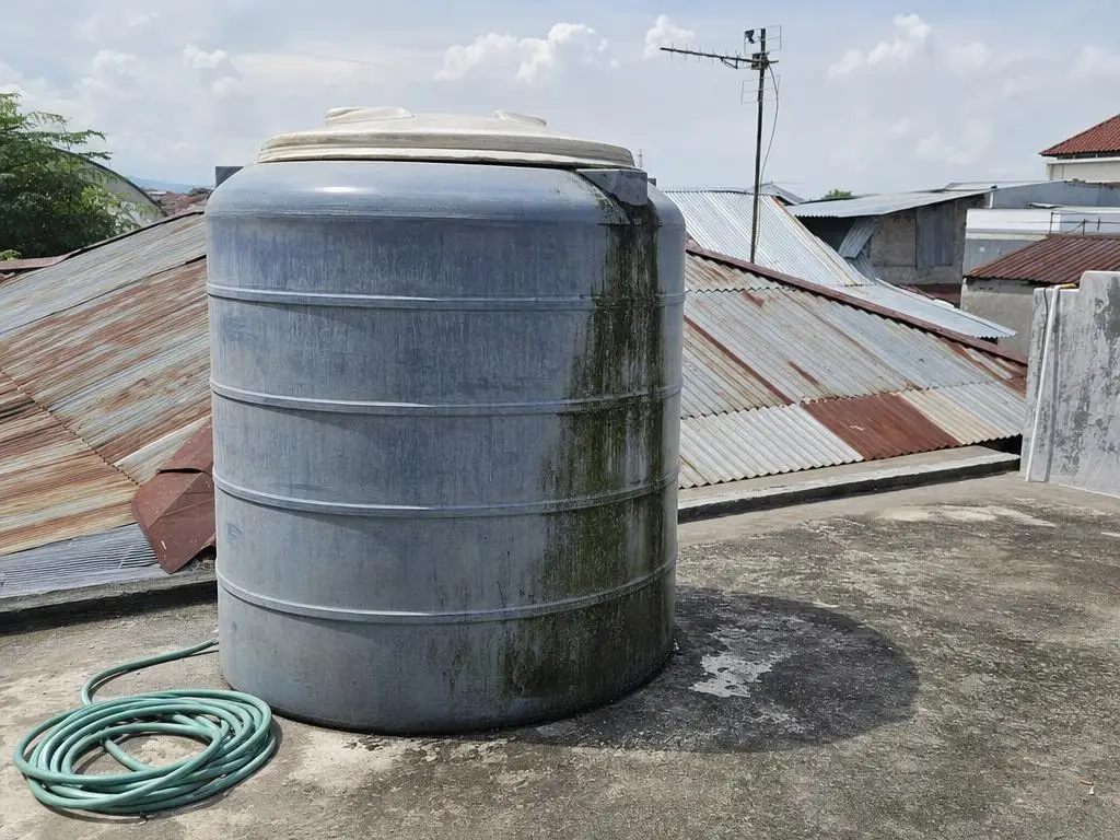 Toren air plastik biru abu abu di dak beton, permukaan luar pudar dengan aliran lumut hijau