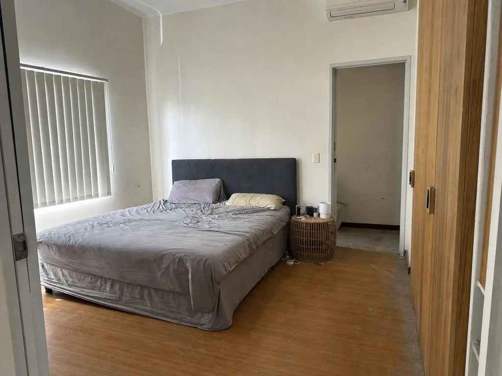 Kamar tidur berdinding putih hangat dengan lantai laminasi kayu, lemari pakaian di sisi kanan, meja rotan bundar di samping ranjang